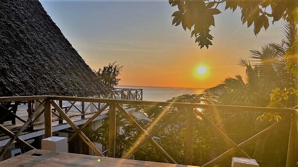 A wooden deck with a thatched roof, overlooking a sunset over the ocean.