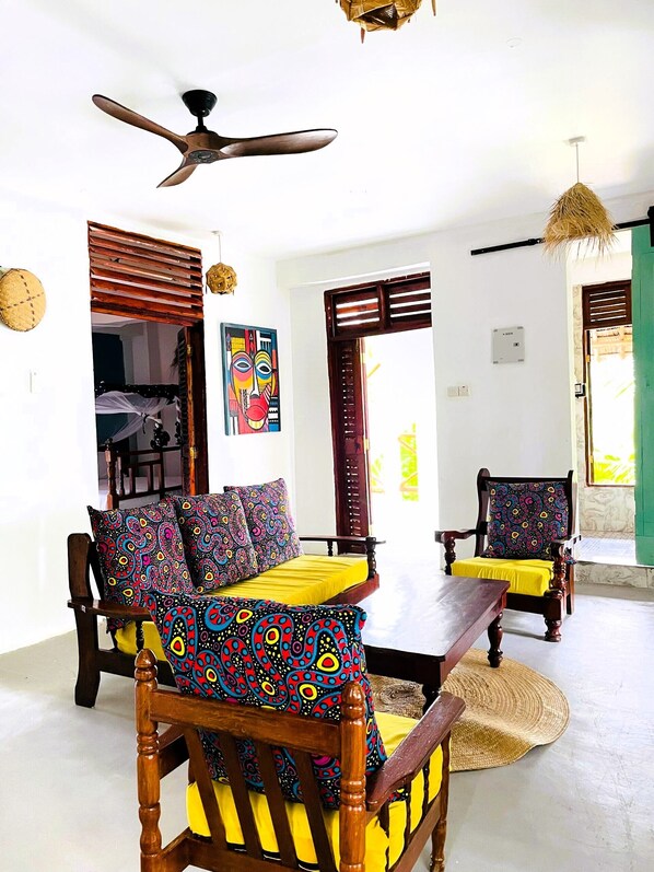 Una habitación con ventilador de techo, un sofá con cojines de colores, una mesa de madera y una silla con un cojín estampado.