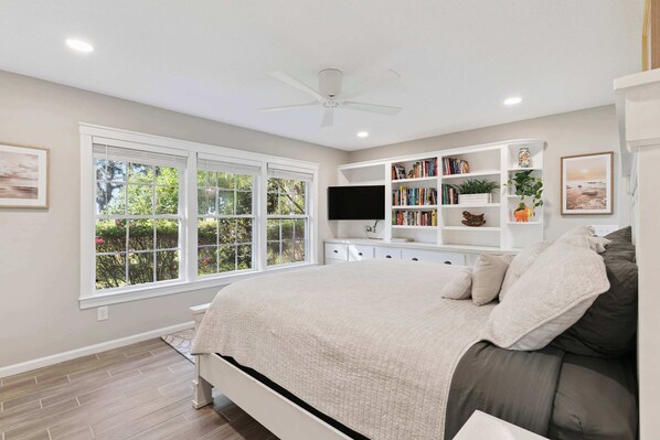 A bedroom with a bed, a ceiling fan, a TV, a bookshelf, and a window with a view of greenery.