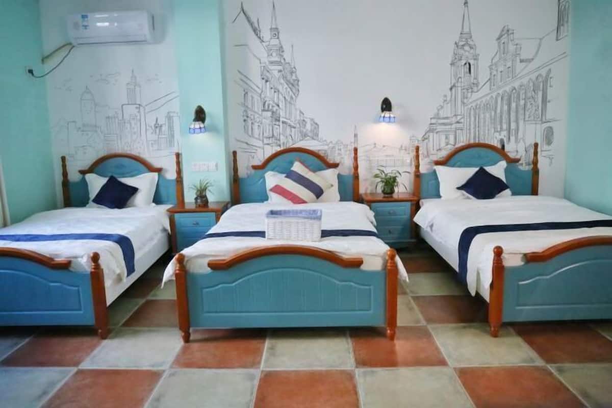 A room with three beds, each with a wooden headboard and a blue mattress cover. There are wall-mounted air conditioning units and a tiled floor.