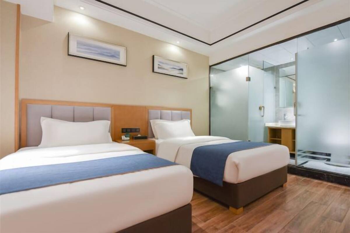 Ein Hotelzimmer mit zwei Betten, einem Holzkopfstück, einem Nachttisch und einer Glastrennwand, die den Raum vom Badezimmer abtrennt.