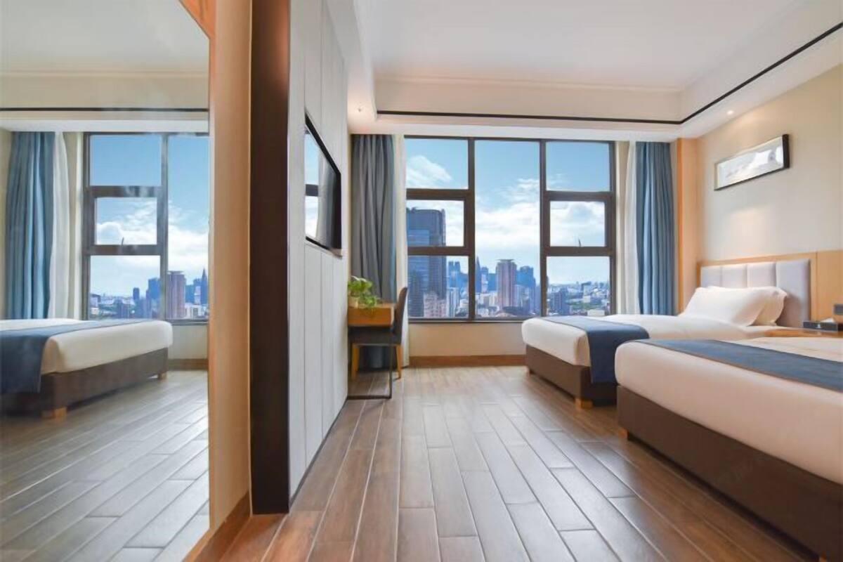 Ein Hotelzimmer mit zwei Betten, einem Schreibtisch und großen Fenstern mit Blick auf die Stadt.