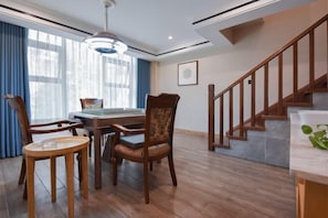 Un comedor con mesa y sillas de madera, una escalera con barandilla de madera y un amplio ventanal con cortinas azules.