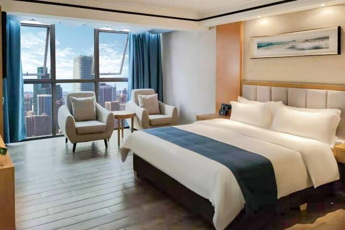 Ein Hotelzimmer mit einem großen Bett, zwei Sesseln, einem kleinen Tisch und Blick auf die Stadt durch große Fenster.