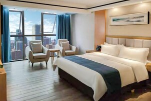 Habitación de hotel con una cama grande, dos sillones, una mesita y vista a la ciudad a través de amplias ventanas.