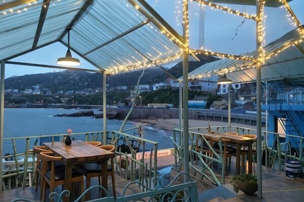 A covered outdoor seating area with tables and chairs, overlooking a coastal view.