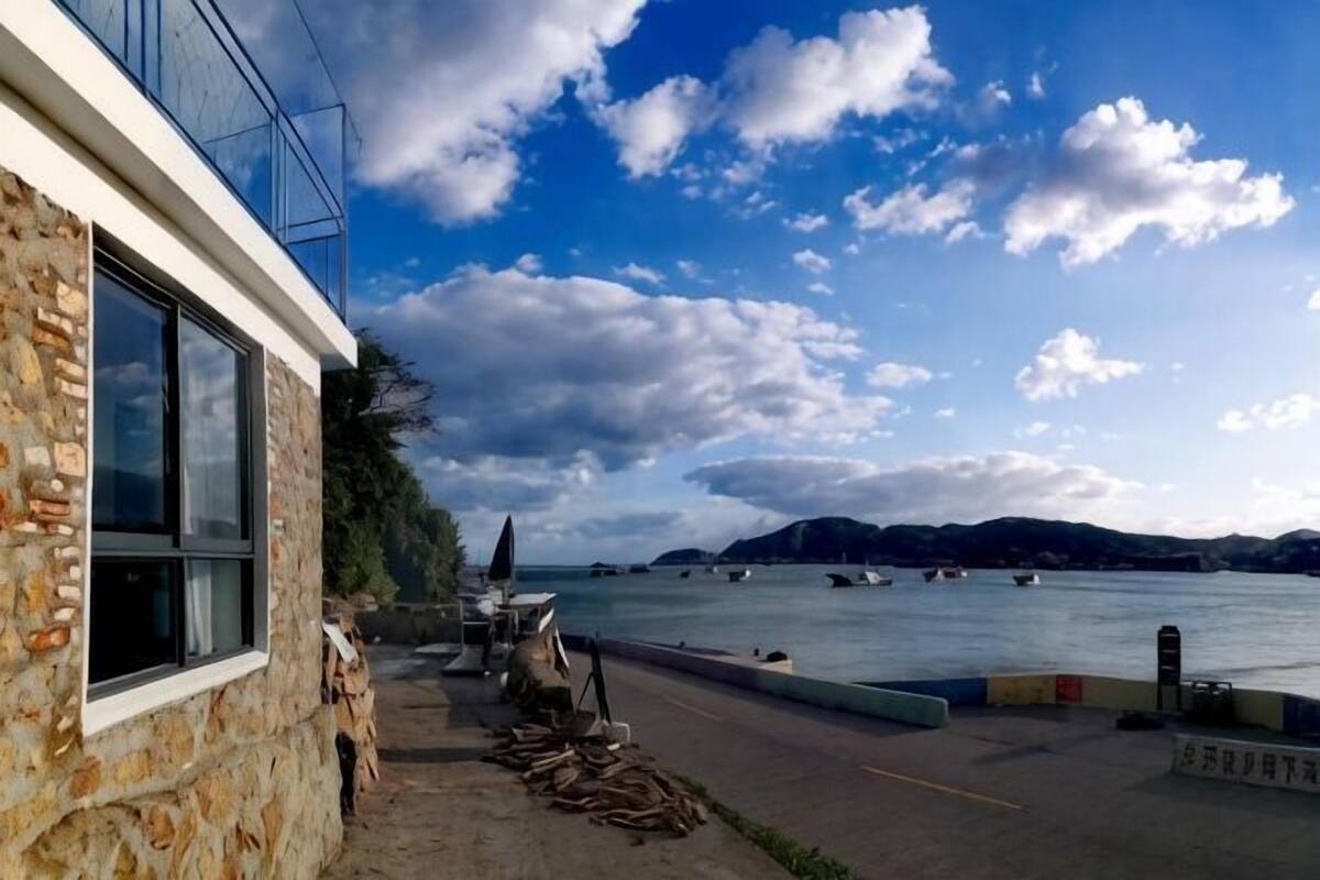 A stone building with a window, a paved area with chairs and a table, and a view of the sea with boats and hills in the background.