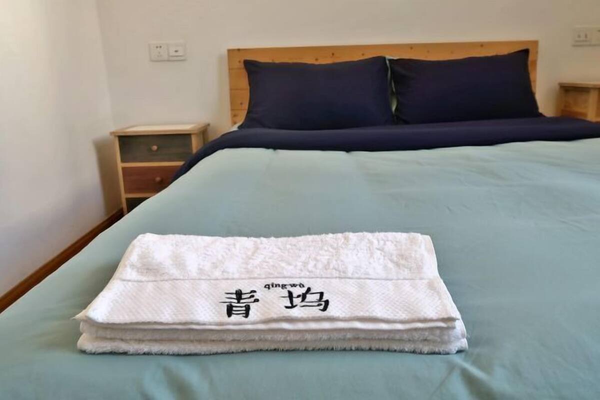 A neatly made bed with a blue bedspread and two pillows, a small wooden nightstand with a drawer, and a folded white towel with Chinese characters on it.
