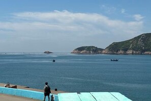 A coastal scene with a person standing on a platform overlooking calm waters, boats in the distance, and a hilly landscape. - Qingwu Fisherman's Hostel (Shengsi)