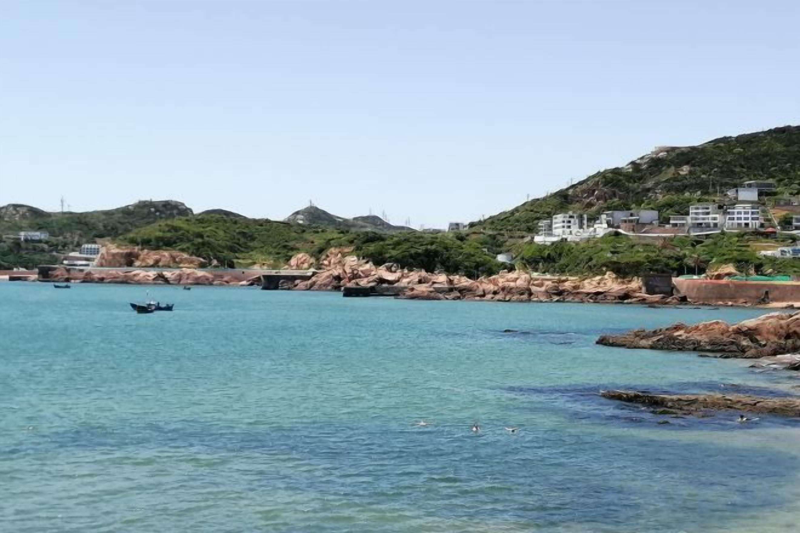 A coastal area with clear water, rocky shoreline, and buildings on a hillside.