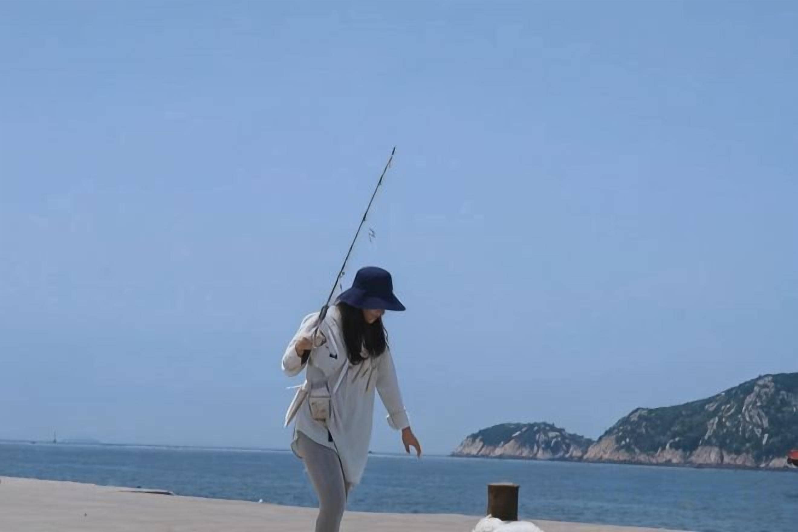 A person fishing by the sea with a clear sky and distant hills.