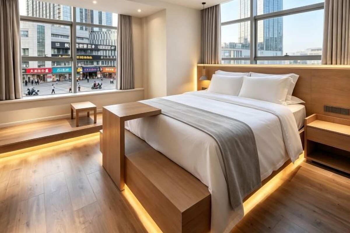 A modern hotel room with a large bed, wooden bedside tables, and a view of a cityscape through large windows.