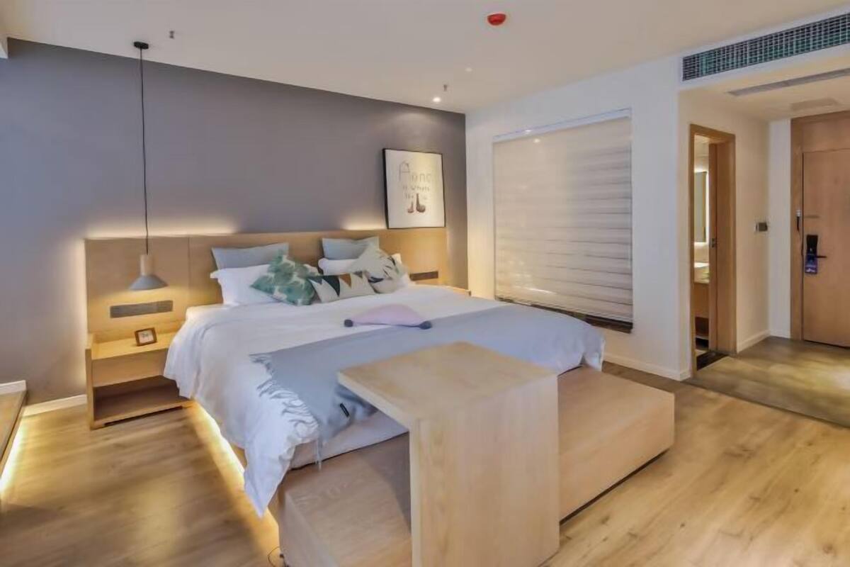A modern bedroom with a bed, bedside tables, a bench, a wall-mounted light, and a window with blinds.