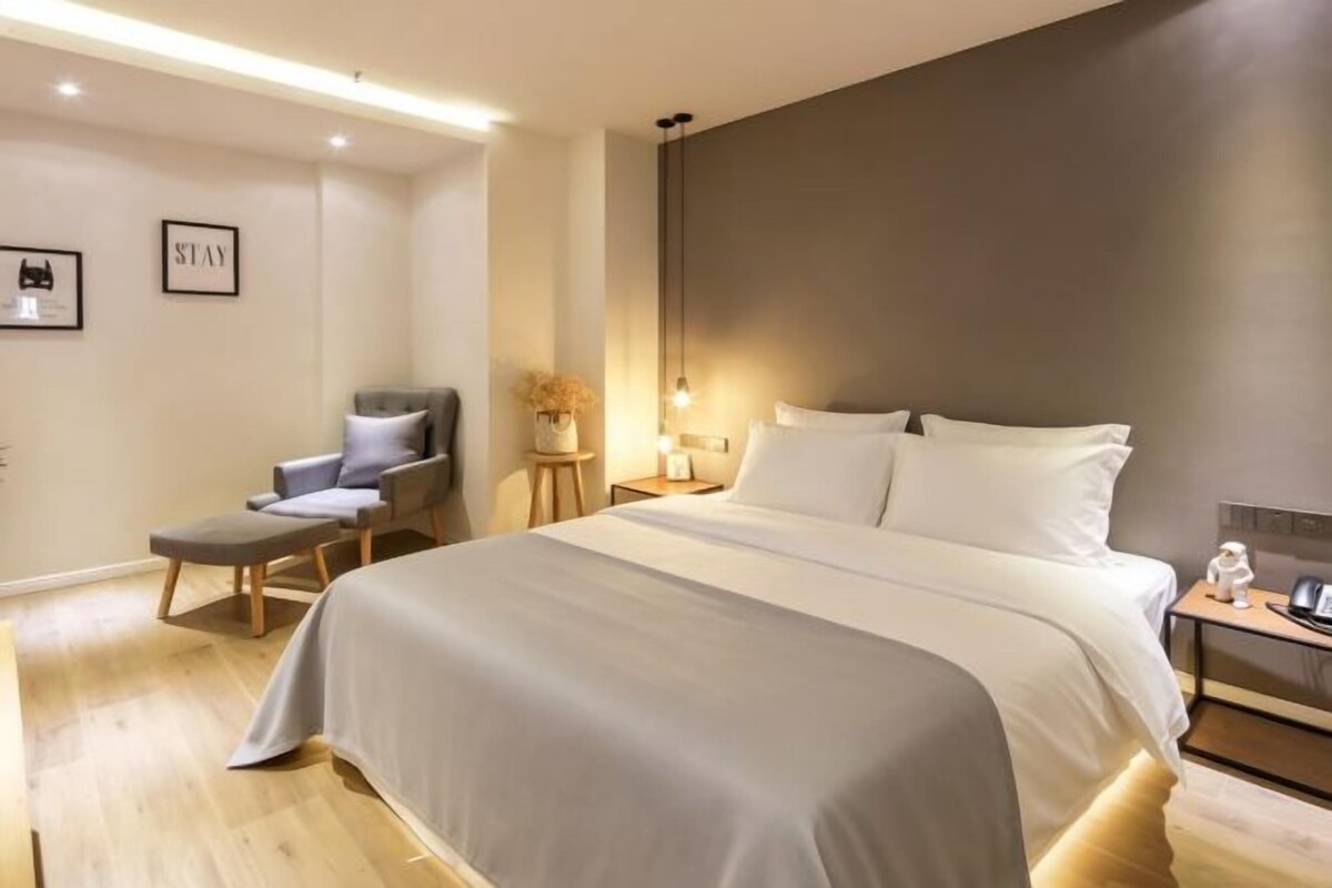 A modern hotel room with a large bed, a seating area with a chair, a bedside table, and a nightstand.