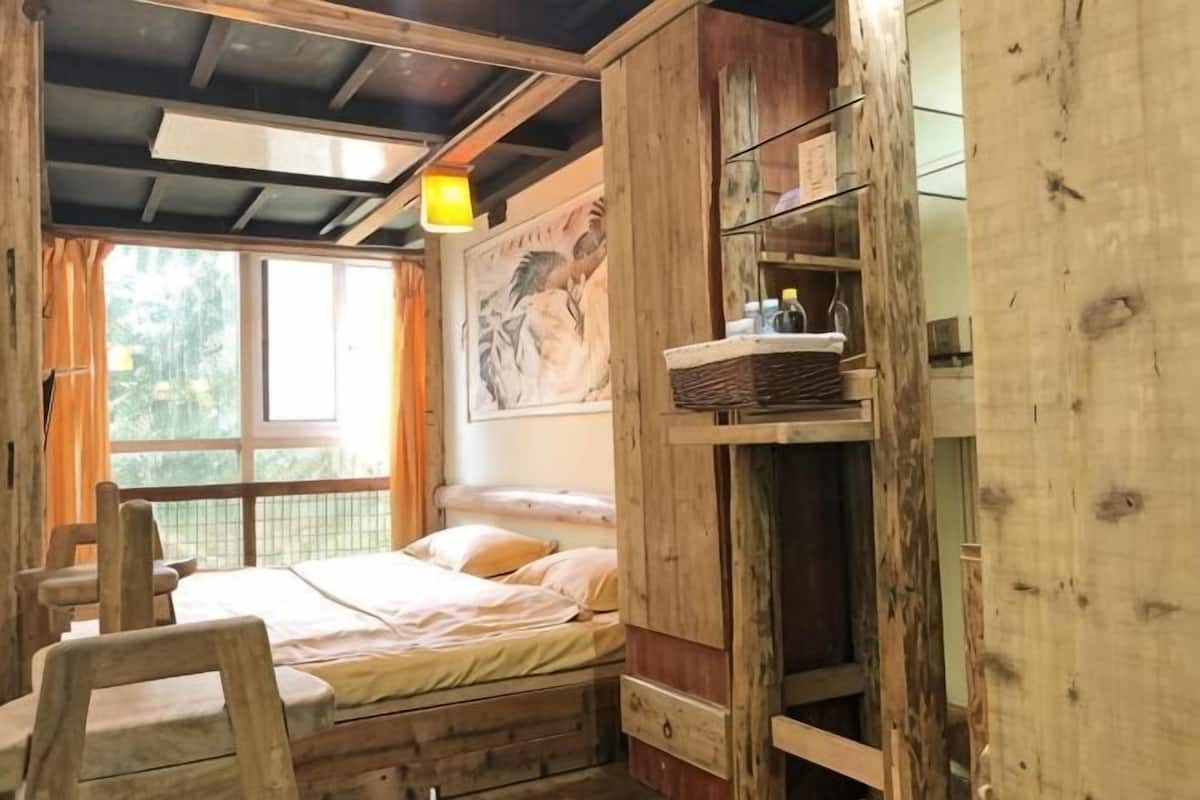 A rustic wooden room with a bunk bed, a wooden chair, a shelf with items, and a window with curtains.