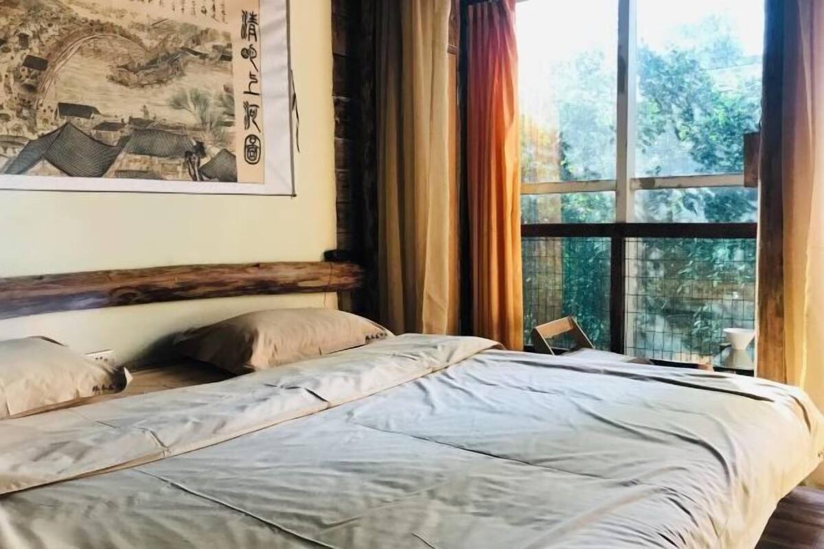 A neatly made bed with a headboard, a window with curtains, and a traditional painting on the wall.