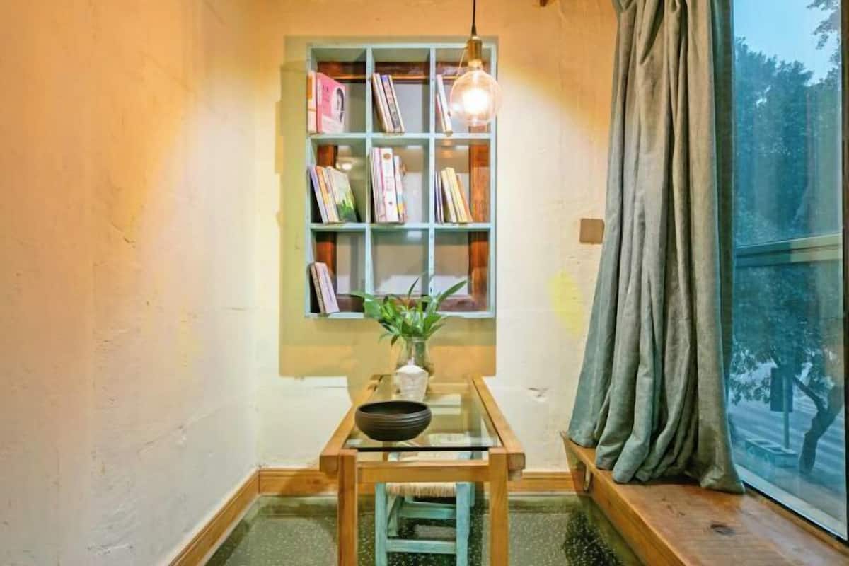 A cozy room with a bookshelf, a small table with a bowl, a hanging light, and a window with curtains.