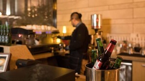Angolo bar con bottiglie in un contenitore metallico, un barista sullo sfondo e un muro in mattoni.