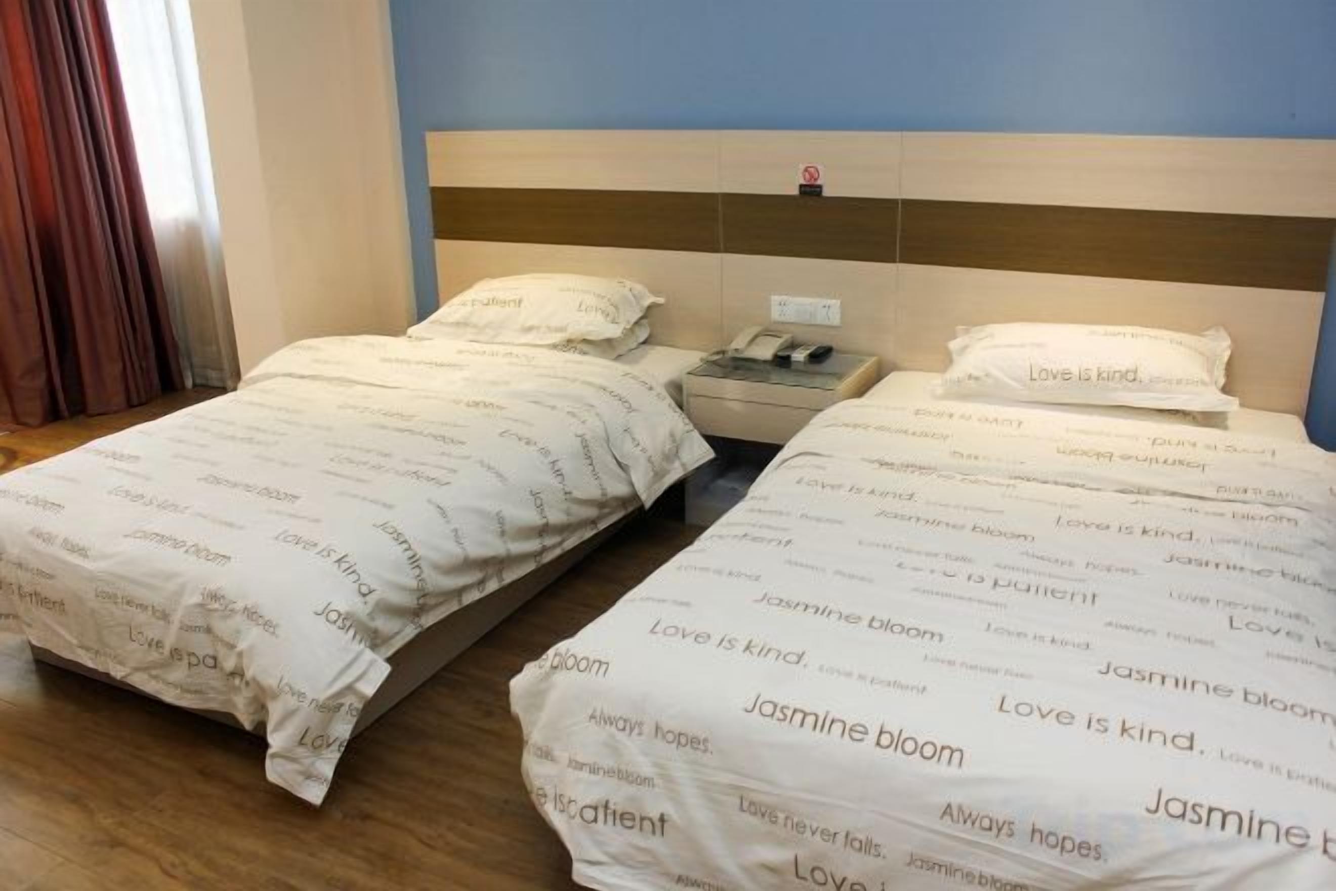 Habitación de hotel con dos camas, cada una con cubrecama blanco con texto impreso. Hay una mesita de noche con una lámpara y un teléfono.