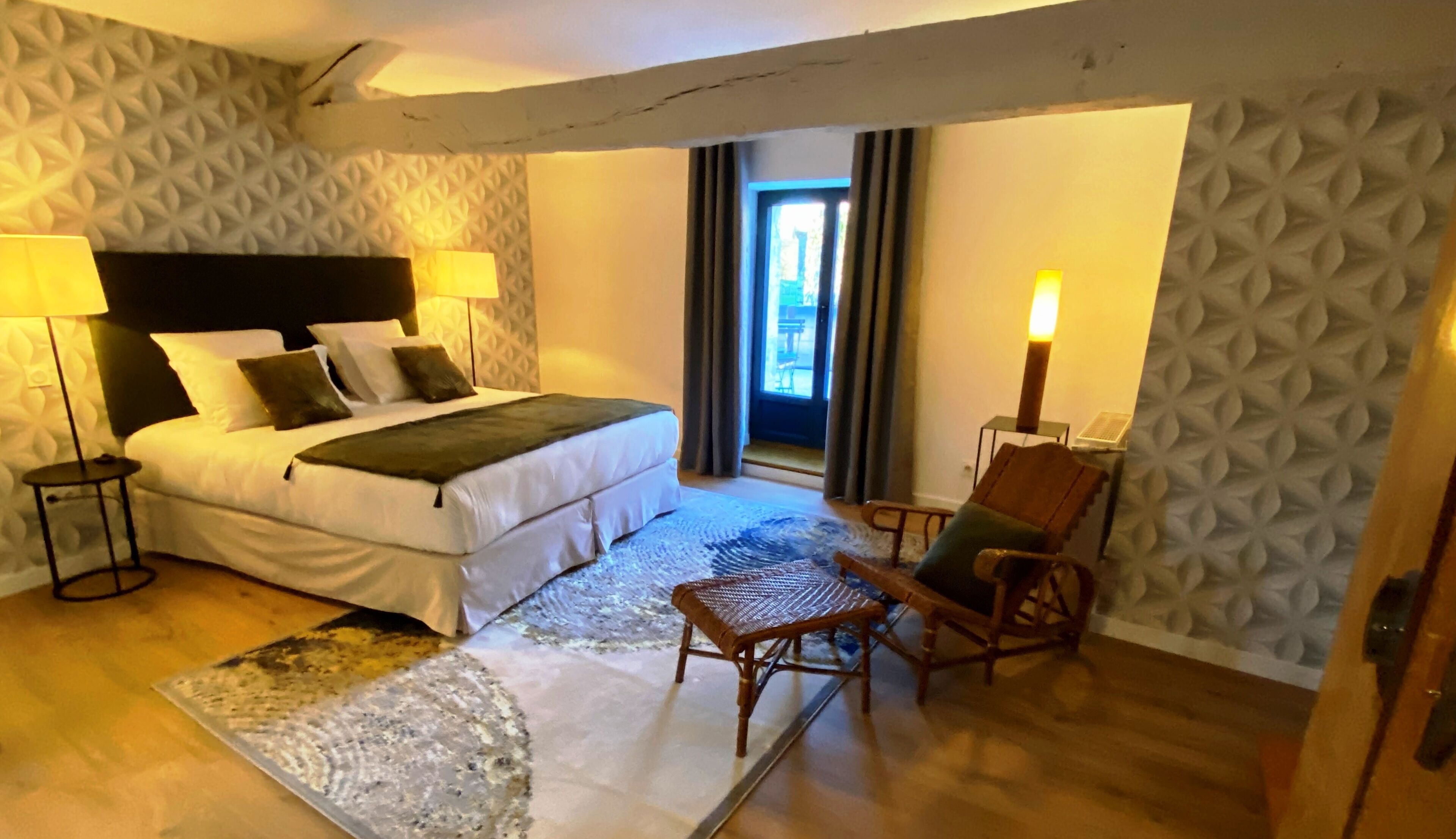 Quarto de hotel com uma cama, duas mesinhas de cabeceira, uma cadeira, uma mesinha, um tapete estampado e uma janela com cortinas.