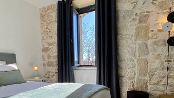 Um quarto com parede de pedra, uma cama com cabeceira, uma janela com cortinas e um abajur.