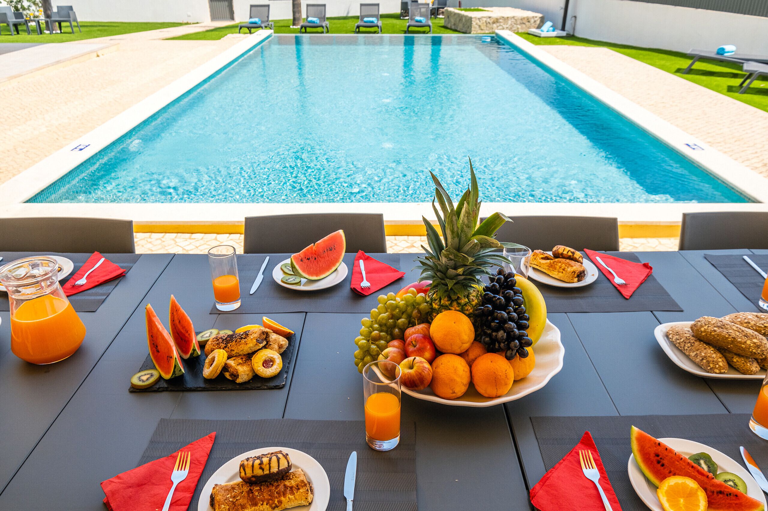 Una mesa de comedor con alimentos para el desayuno, un frutero y vista a una piscina.