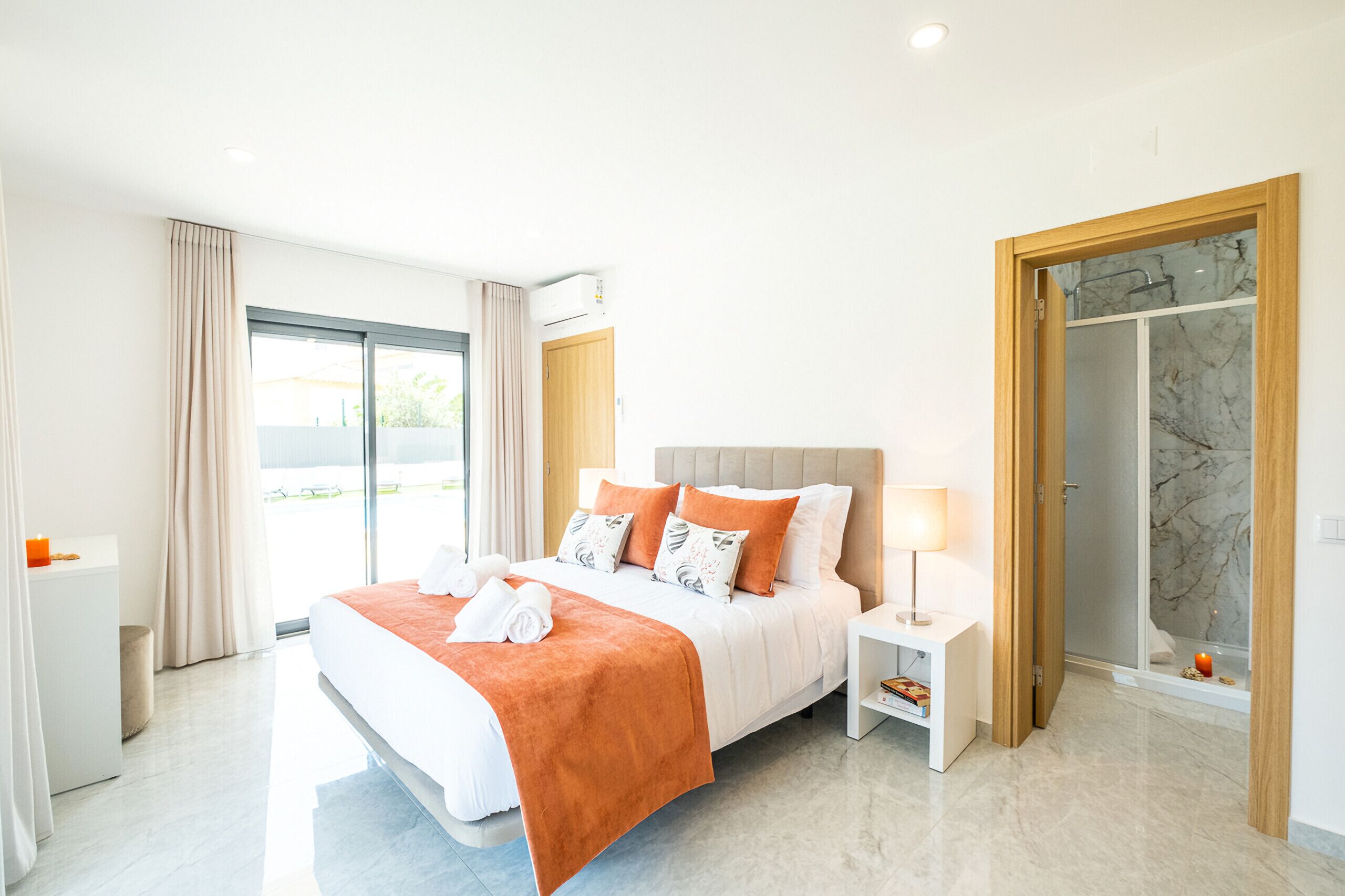 Habitación de hotel moderna con una cama grande, una mesita de noche, un baño con ducha de vidrio y vistas al exterior.