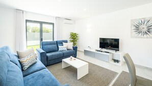 A modern living room with a blue sofa, a white coffee table, a TV on a white cabinet, and a large window. - Family villa with heatable pool and BBQ (Albufeira)