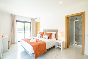 Una habitación de hotel moderna con una cama grande, una mesita de noche con lámpara, un box de ducha de vidrio y vistas al exterior.