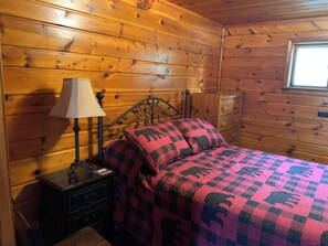 Ein Zimmer in einer Holzhütte mit einem Bett, einem Nachttisch, einer Lampe und einem Fenster.