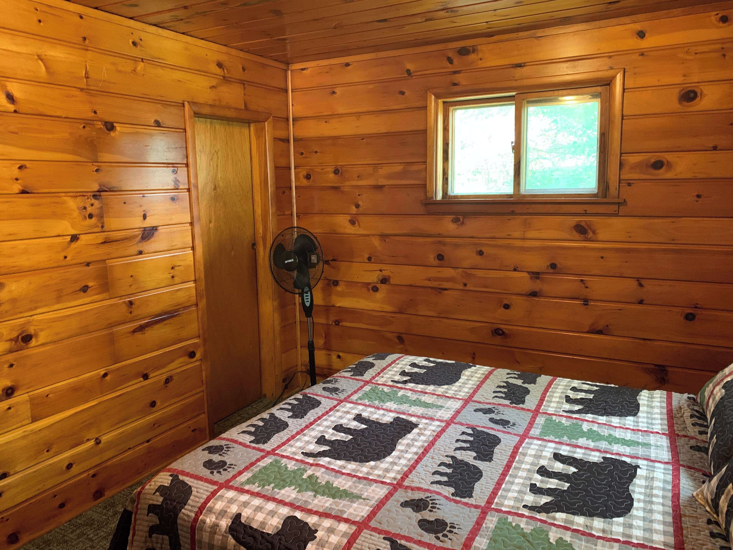 Una habitación de cabaña de madera con una cama, una ventana y un ventilador fijado a la pared.