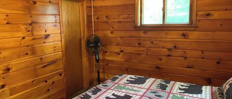Een houten kamer met een bed, een raam en een ventilator aan de muur.