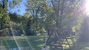 A playground with swings and a merry-go-round in a grassy area with trees in the background. - Gresham Shores - Hungry Bear - PET FRIENDLY (Arbor Vitae)