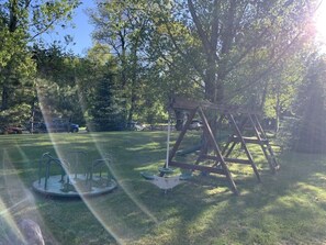Ein Spielplatz mit Schaukeln und Karussell in einer grünen Wiese, im Hintergrund Bäume.