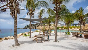 A beachside area with tables and chairs under palm trees, a pool, and a view of the sea. - Blue Bay Green View (Sint Michiel)