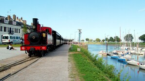 Une locomotive à vapeur sur des rails, à côté d’un canal où se trouvent des bateaux.
