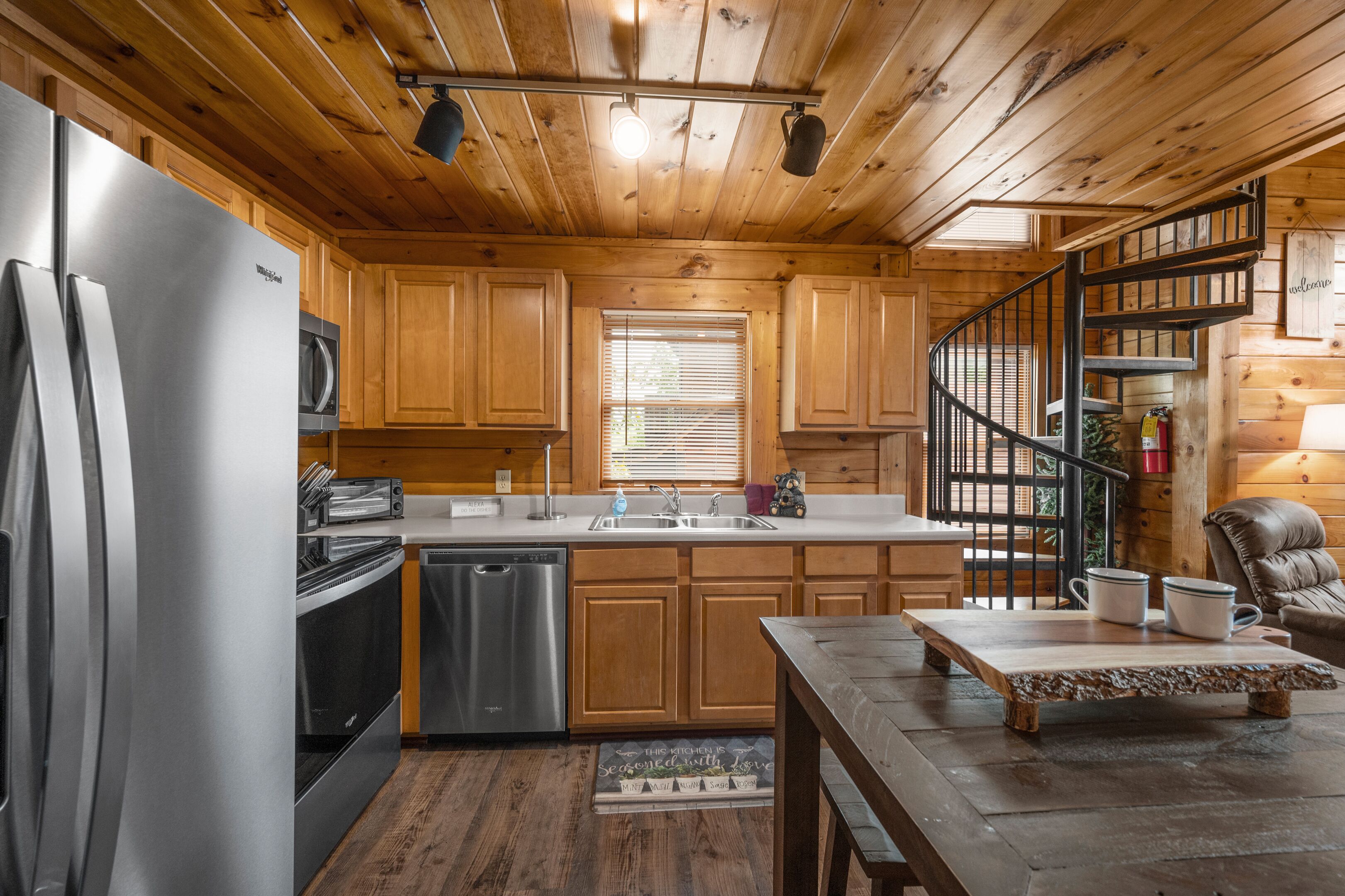 A wooden cabin kitchen with a stainless steel refrigerator, microwave, and oven. There is a wooden dining table with two cups and a bowl. A spiral staircase leads to a loft area.