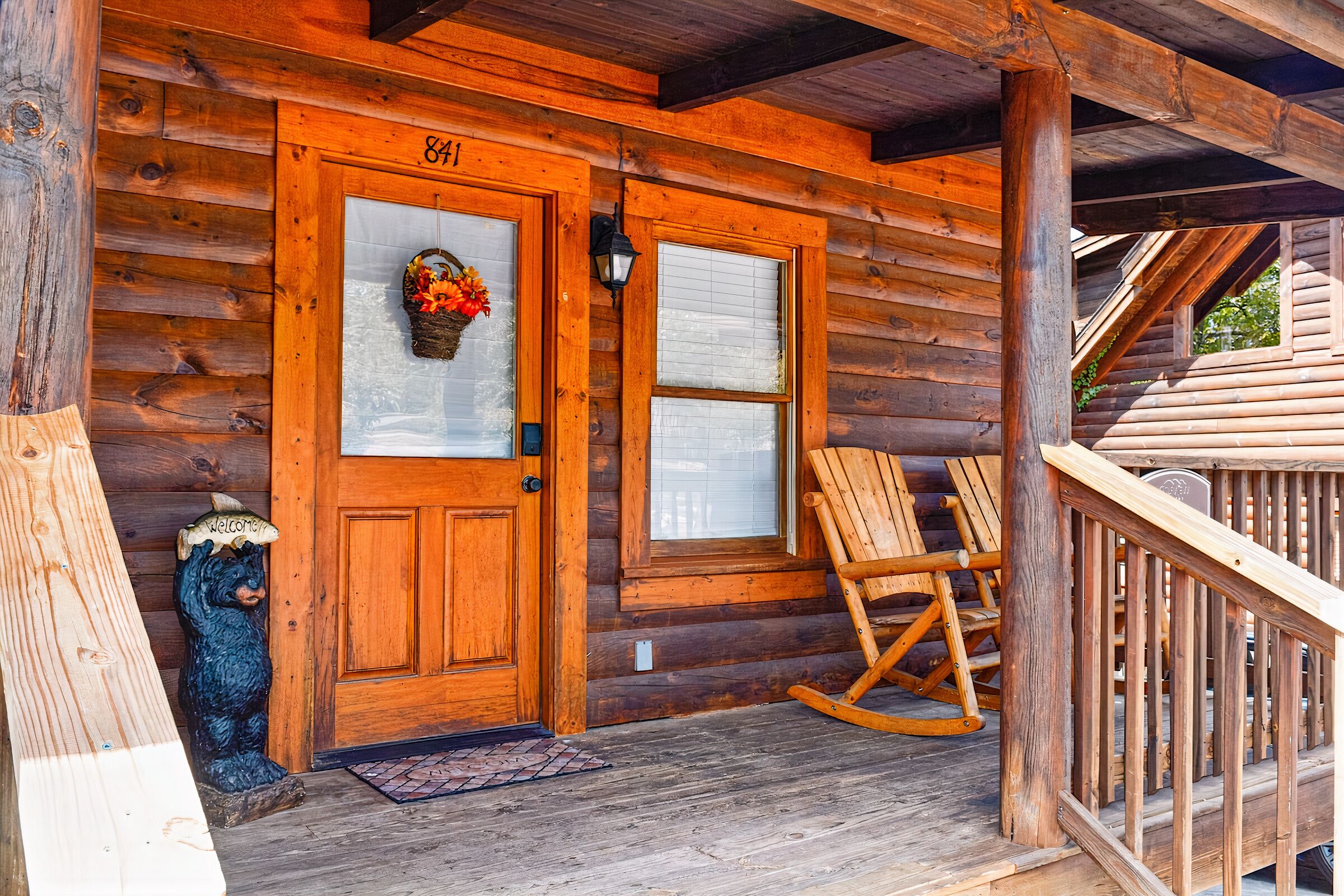 A wooden cabin with a double door, a welcome bear statue, a rocking chair, and a porch with a railing.