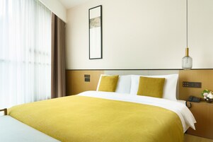 Habitación de hotel con una cama grande, una mesita de noche con teléfono y un florero, una lámpara de pared y una ventana con cortinas.
