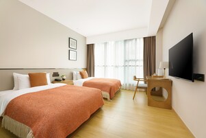 A hotel room with two beds, a TV, a desk, and a large window with curtains.