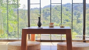 A room with a wooden table, two round woven ottomans, and a view of a forest through large windows. - Blossom Hill Hotel Ya'an Mengdingshan (Ya'an)