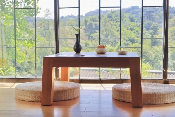 A room with a wooden table, two round woven ottomans, and a view of a forest through large windows. - Blossom Hill Hotel Ya'an Mengdingshan (Ya'an)