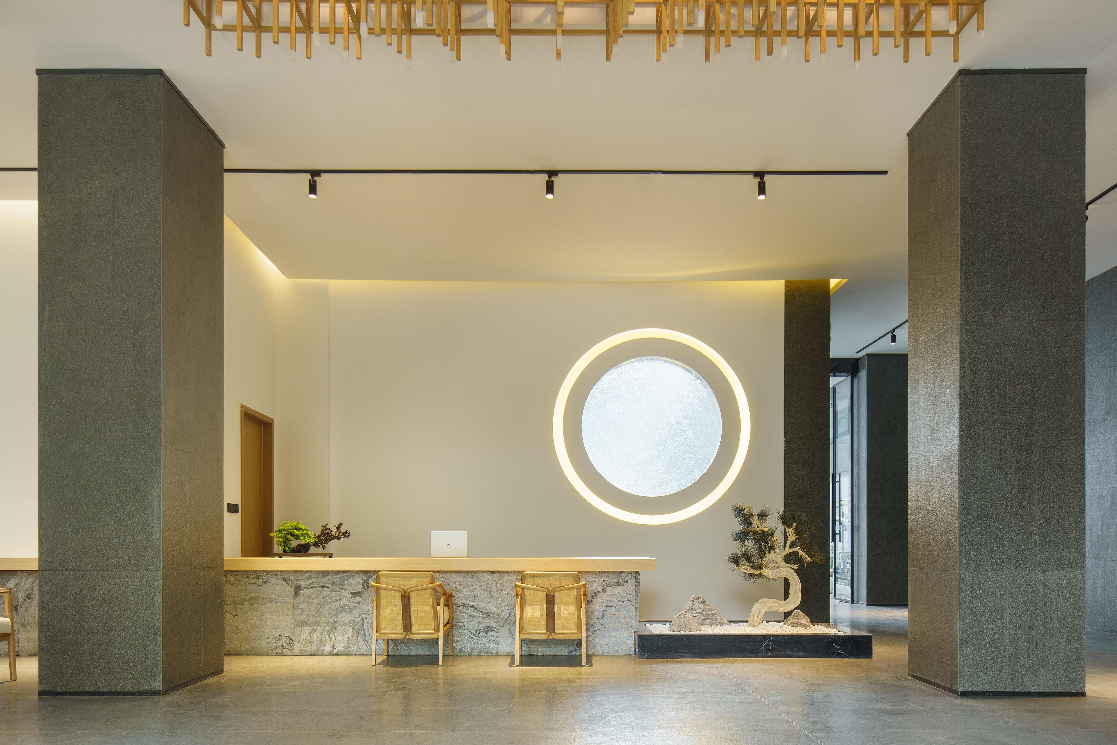 A modern reception area with a marble counter, two wooden chairs, a round illuminated mirror, and a potted plant.