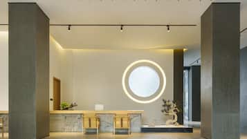 A modern reception area with a marble counter, two wooden chairs, a round illuminated mirror, and a potted plant.