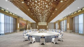 Un amplio salón de conferencias con una mesa redonda rodeada de sillas blancas con lazadas azules, techo de madera con luces colgantes y grandes ventanales a los lados.