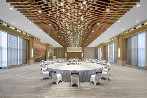 A large conference room with a round table surrounded by white chairs with blue sashes, a wooden ceiling with hanging lights, and large windows on the sides.
