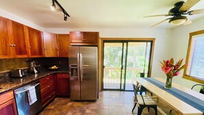 A kitchen with wooden cabinets, a stainless steel refrigerator, a dining table with chairs, and a view of the outdoors through a sliding glass door. - Fresh, Bright, Cute. Near Hanalei! Walk to the ocean in minutes. (Princeville)