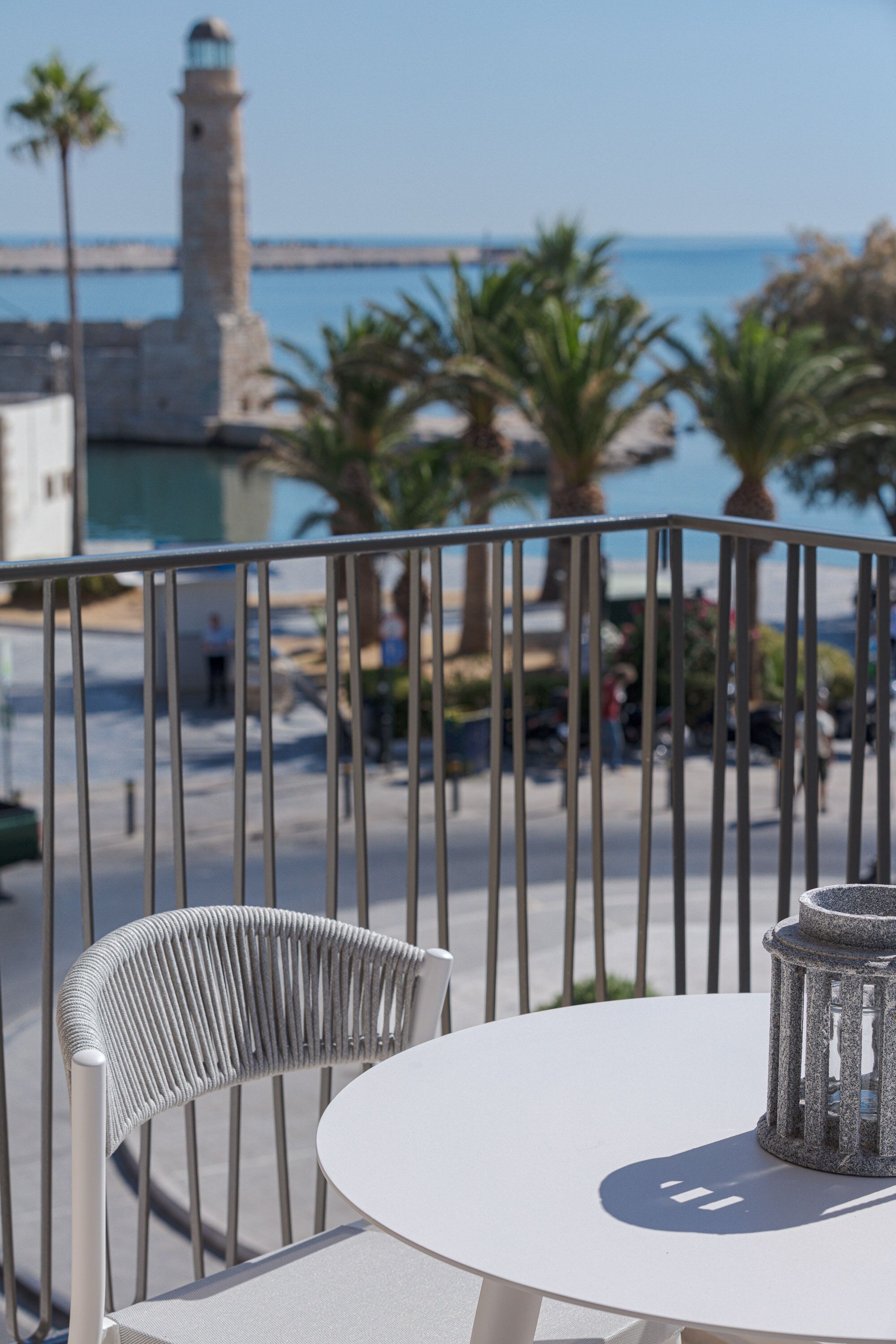 A balcony with a white table and chair, overlooking a coastal area with a lighthouse and palm trees.