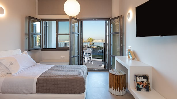 A bedroom with a bed, a TV, a bookshelf, and a balcony with a view. - Modernly furnished family suite with sea view, just in front of the Palm beach (Rethymno)