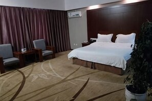 Une chambre d’hôtel avec un grand lit, deux fauteuils, une petite table, une plante en pot et une fenêtre avec des rideaux.